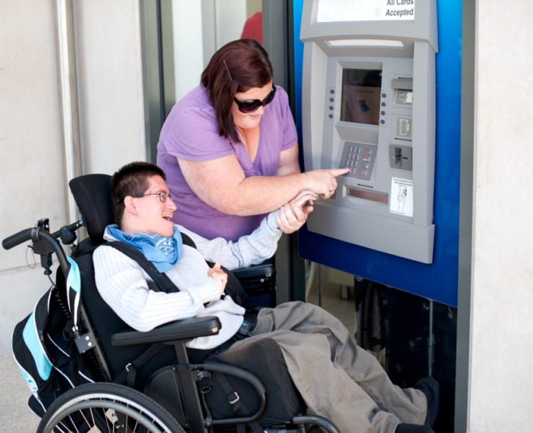 A support worker helps a community member in a wheelchair to use an ATM