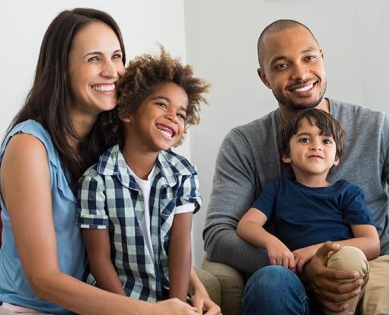A mixed race family sits together on the couch, with a mom, a dad, and two young boys.